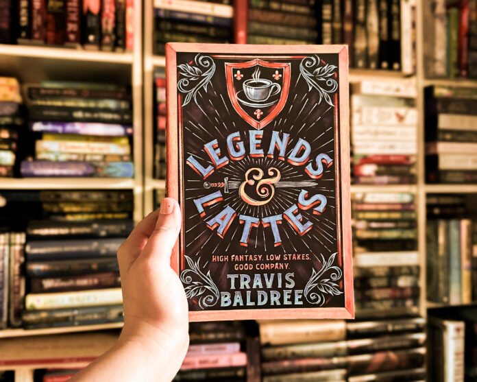 The UK cover of Legends & Lattes by Travis Baldree held up in front of an overflowing bookshelf
